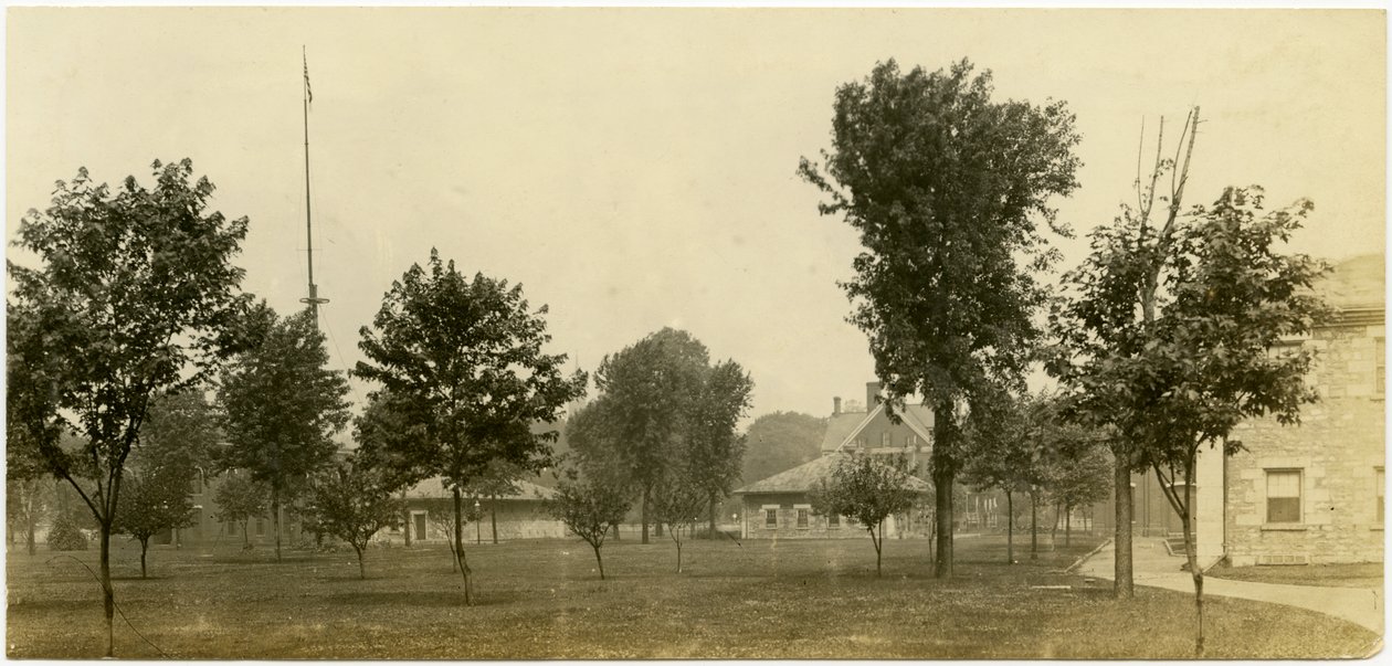 Photographie horizontale sépia montrant un groupe de bâtiments en briques bordant un espace vert ouvert avec des arbres dispersés. Certains des bâtiments en briques semblent être des maisons - Carl Michel