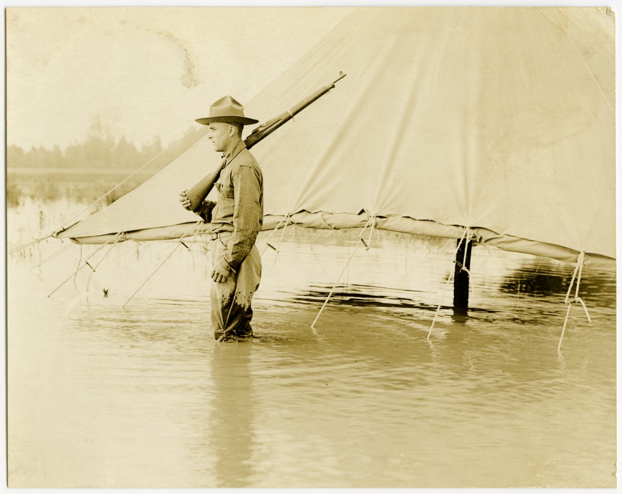 Photographie horizontale sépia montrant un soldat en uniforme monté la garde dans un champ inondé. Le soldat se tient à côté d